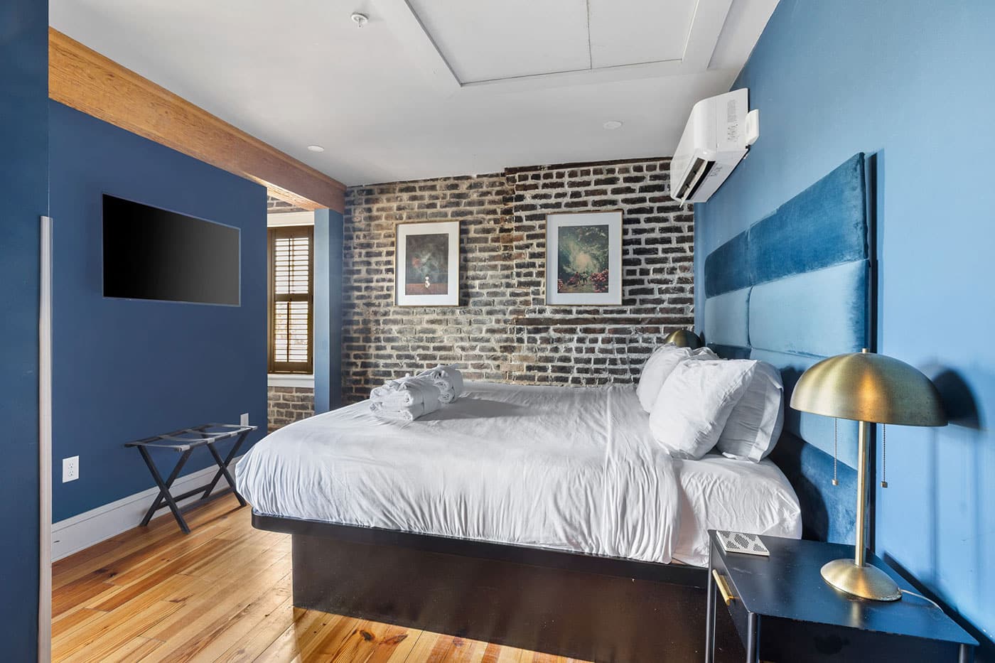 Blue-toned bedroom with a white bed, brick accent wall, two framed pictures, and a wall-mounted TV on the blue wall at left.