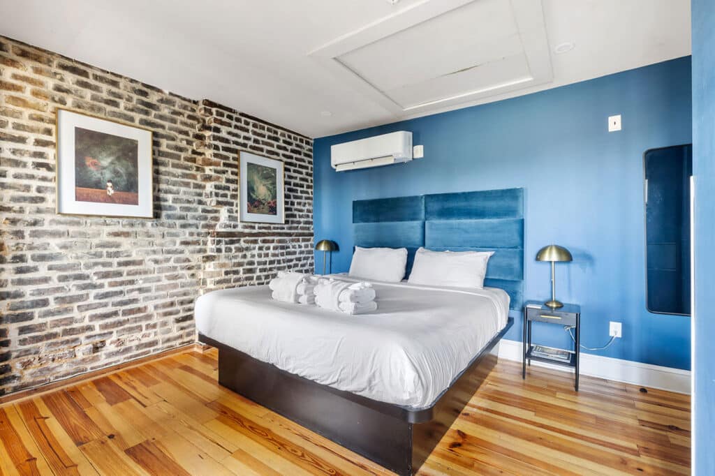 Bedroom with exposed brick wall on left, blue accent wall behind a king bed with white linens and folded towels on top. Dark wood bed frame, two brass bedside lamps, framed artwork, and wooden floor create a modern, cozy look.
