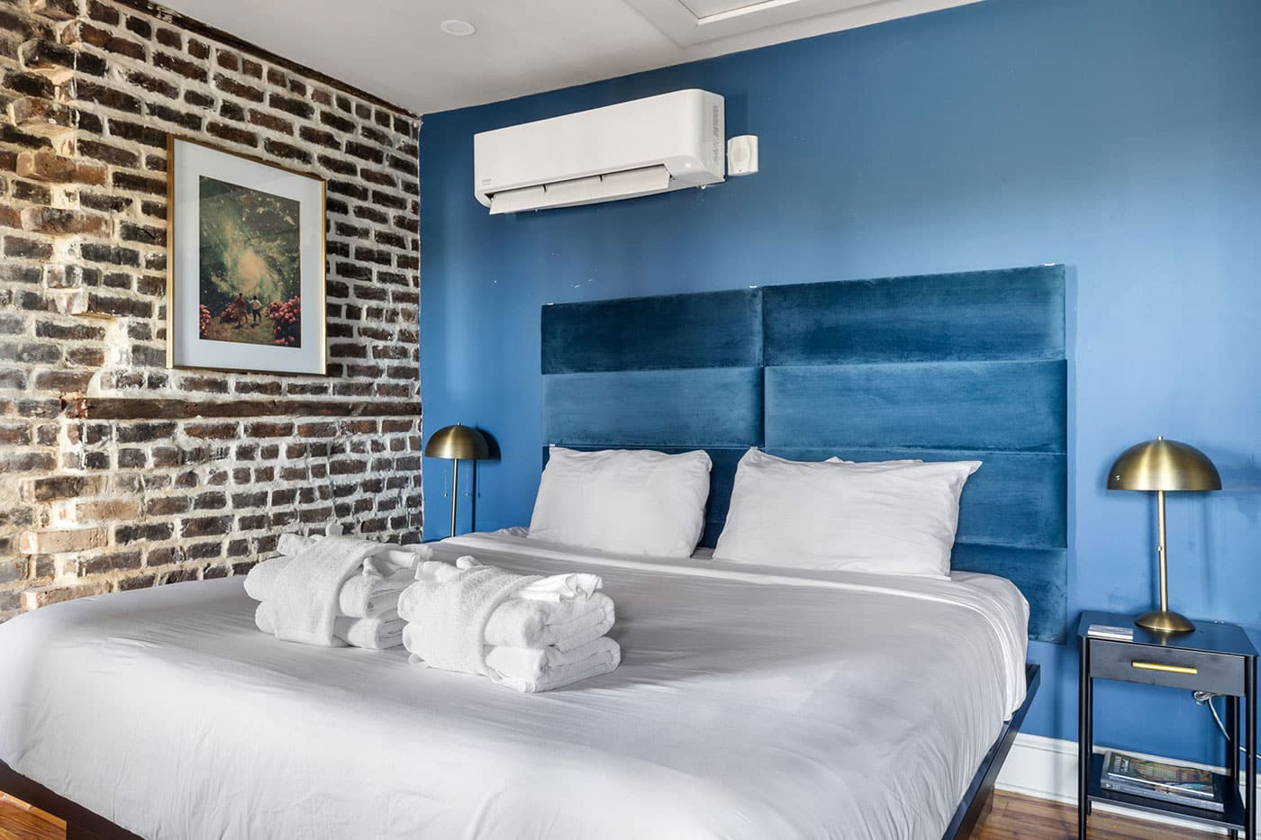 King bed with white linens and a blue upholstered headboard in a blue-walled hotel room with a brick accent wall and brass lamps.