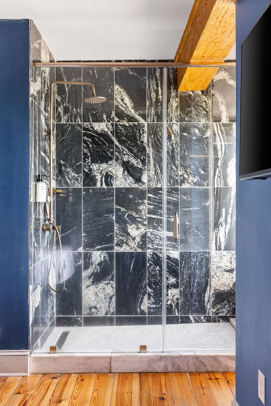 Walk-in shower with a glass door, dark marbled tile walls, brass fixtures, and a wooden beam ceiling against blue walls.