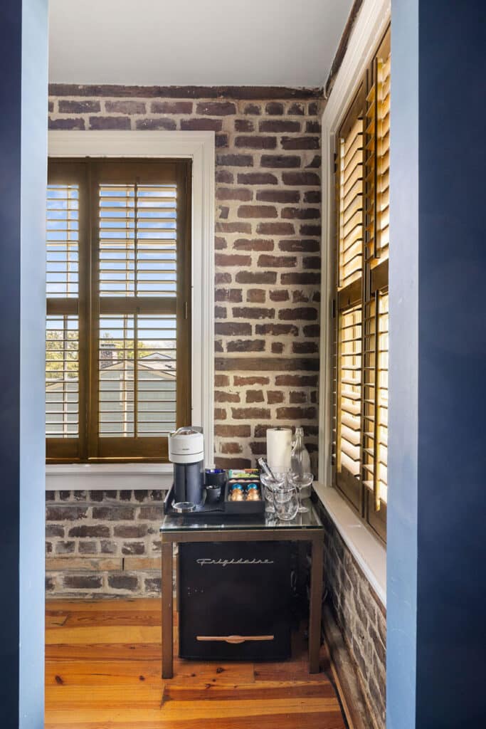 Coffee station in a brick-walled corner: small fridge beneath a glass table, coffee maker, cups, and a carafe next to a window with wooden blinds.