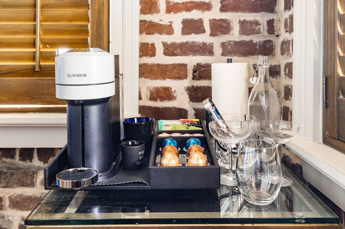 Nespresso coffee machine on a black tray with capsules, cups, and glassware on a glass counter against a brick wall.