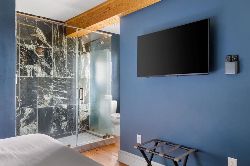 Blue-walled hotel room with a mounted flat-screen TV and a glass-walled shower area featuring black marble tiles. A wooden beam spans the ceiling above. A luggage rack sits nearby. A toilet is visible through the shower door.