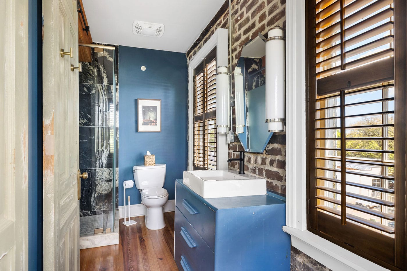 Blue bathroom with brick accent wall, white vessel sink on a blue vanity, toilet, and a glass shower with wooden blinds on the window.