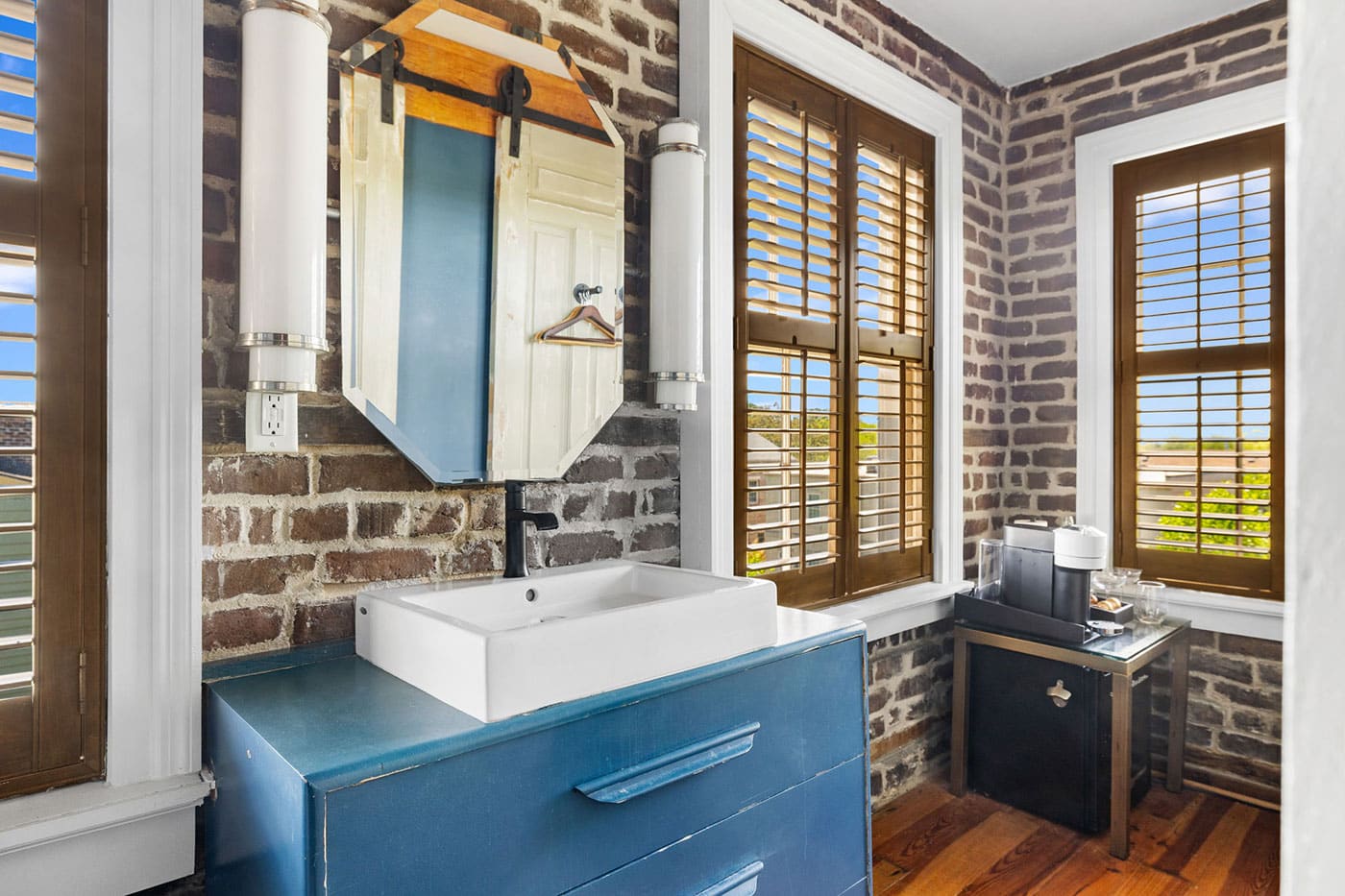 Brick-walled bathroom with a blue vanity, white rectangular sink, and large octagonal mirror on an exposed brick wall with wooden shutters.