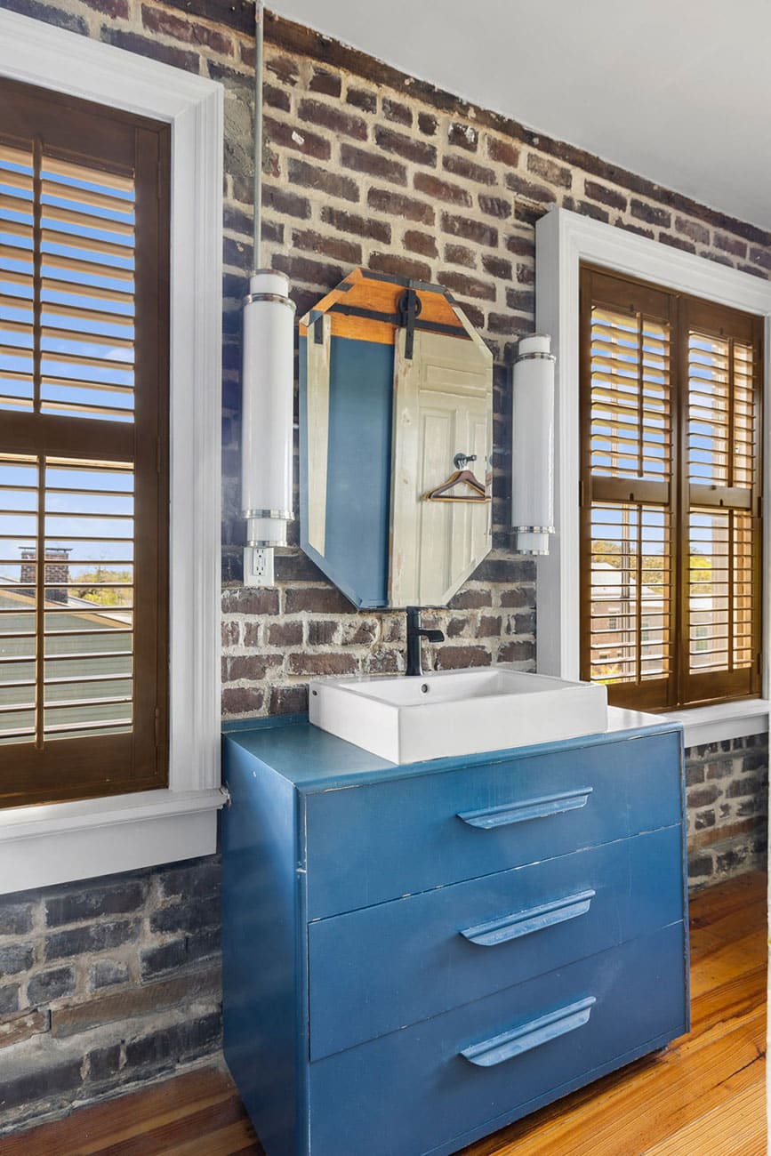 Blue three-drawer vanity with a white square sink and black faucet against a brick wall and wooden-window frames far left and right.