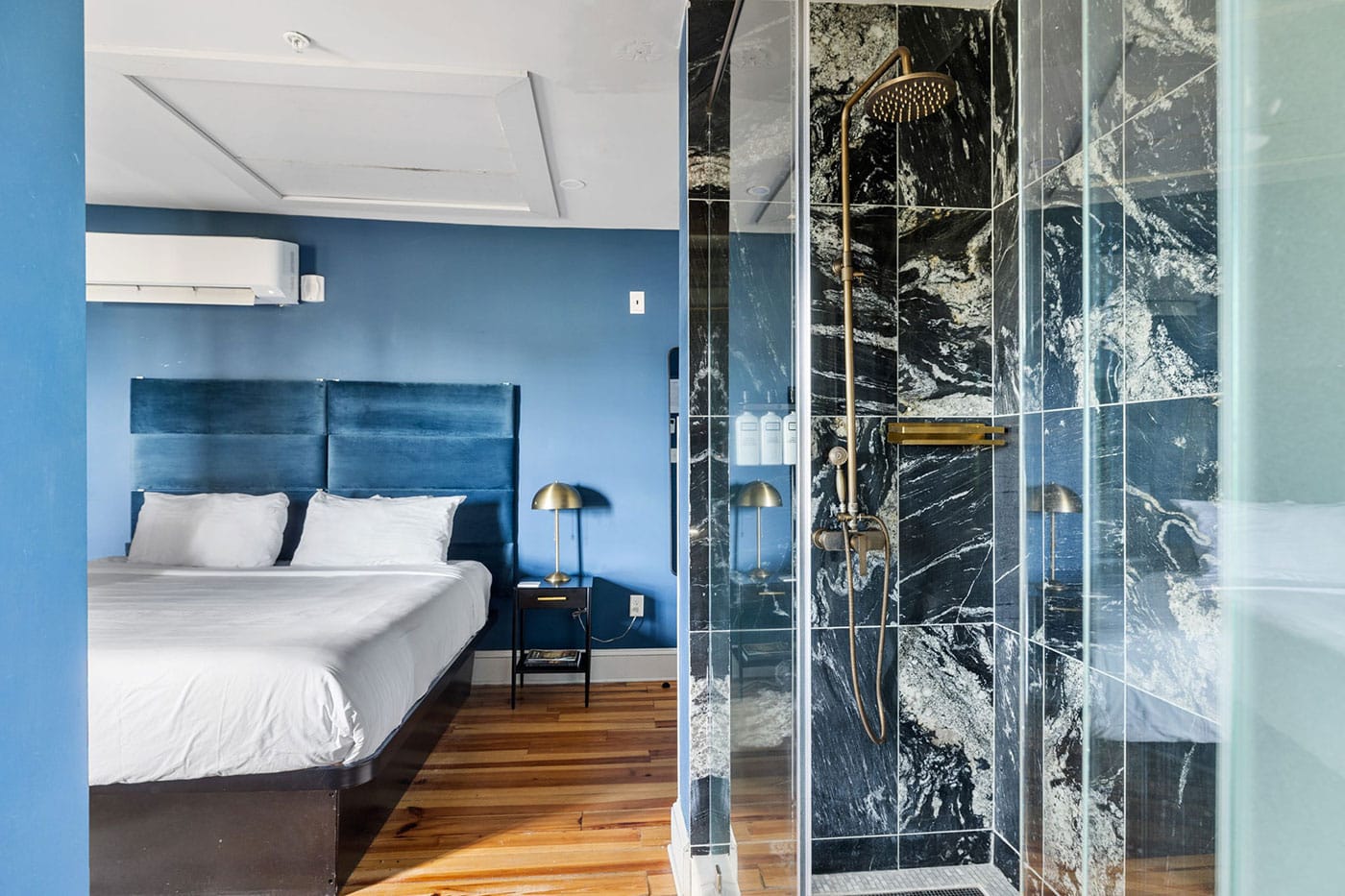 Hotel bedroom with a blue upholstered headboard bed, white pillows, and a brass bedside lamp; glass-walled marble shower on the right.