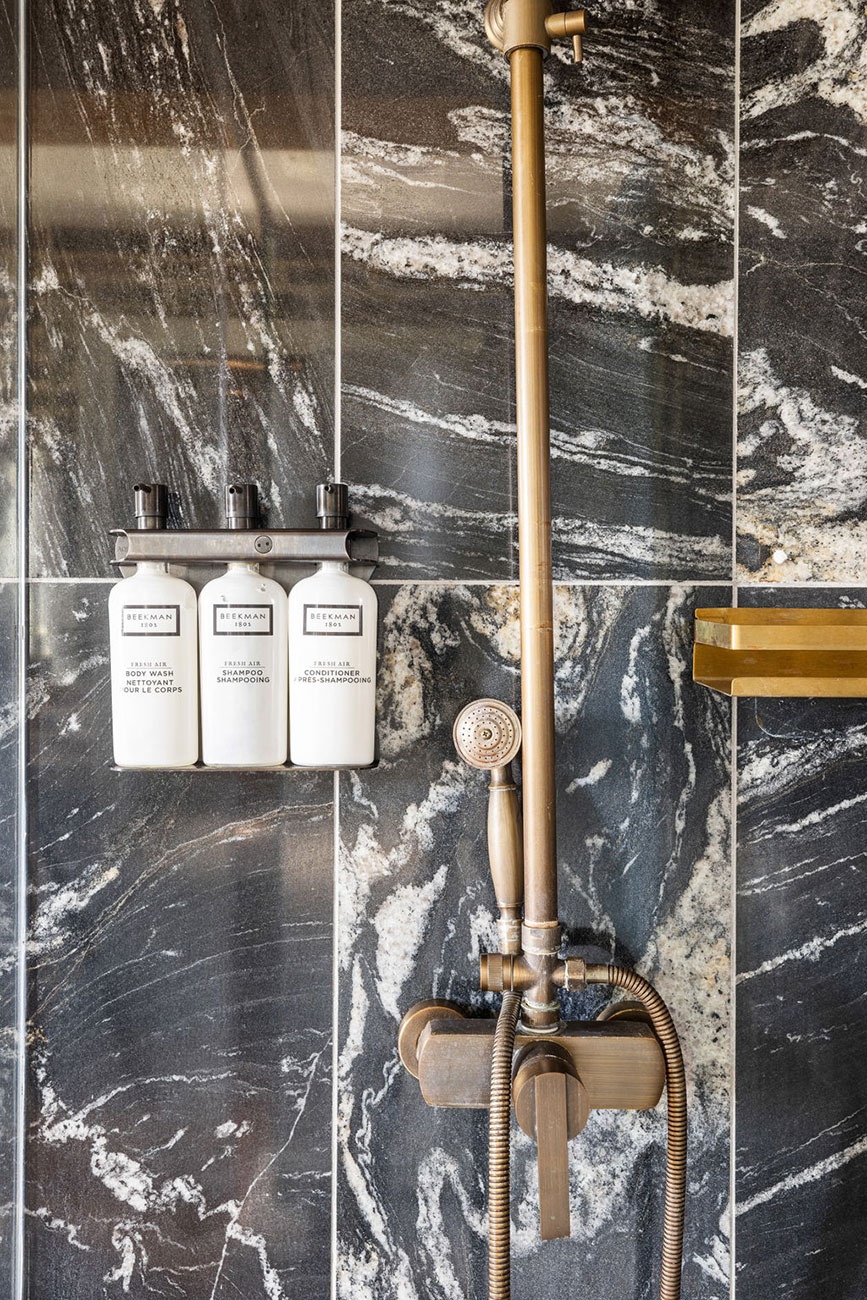 Copper shower fixtures and handheld wand mounted on a dark marble wall, with three white Beekman shampoo/conditioner bottles on a metal shelf.