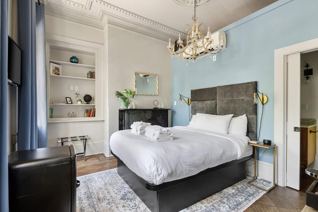 Bedroom with white linens on a platform bed, gray upholstered headboard, and a blue accent wall under a crystal chandelier. Curved curtains and a built-in bookshelf add elegance.