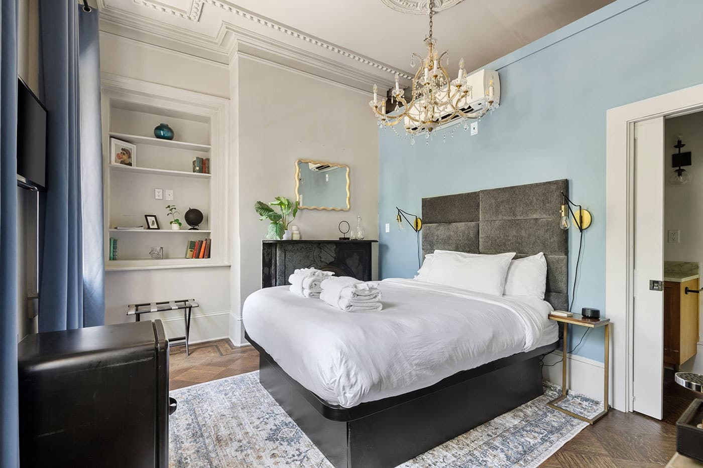 Bedroom with white linens on a platform bed, gray upholstered headboard, and a blue accent wall under a crystal chandelier. Curved curtains and a built-in bookshelf add elegance.