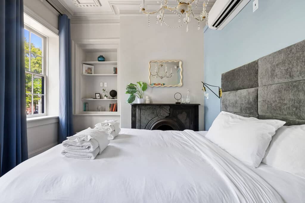 Bright bedroom with white bedding, blue curtains, and a chandelier over a dark mantelpiece and window light.