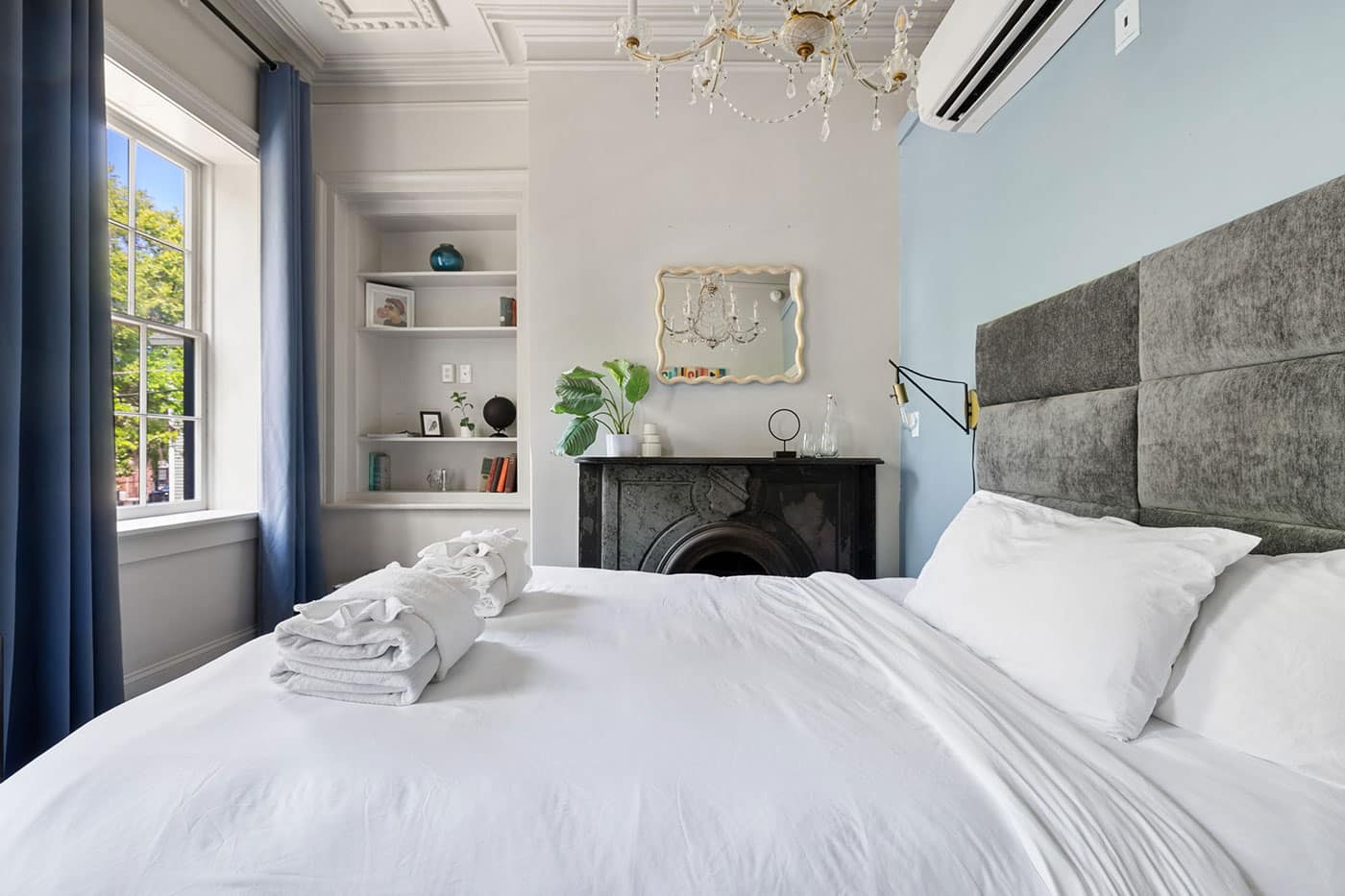 Bright bedroom with white bedding, blue curtains, and a chandelier over a dark mantelpiece and window light.