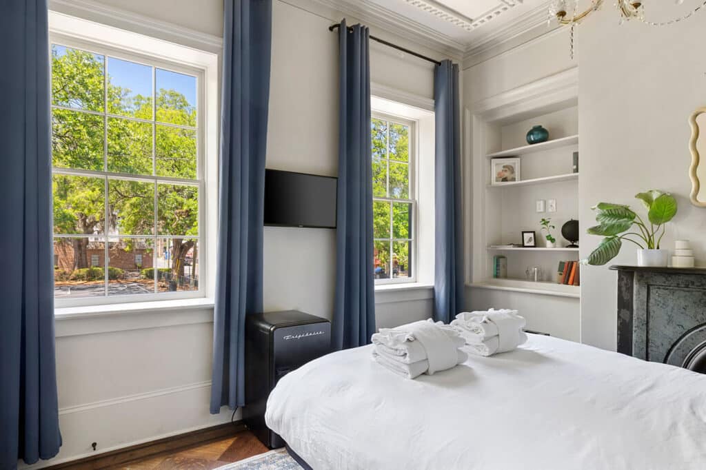 Bright bedroom with large windows framed by blue curtains, a bed with white linens and folded towels, and built-in shelves on the right.