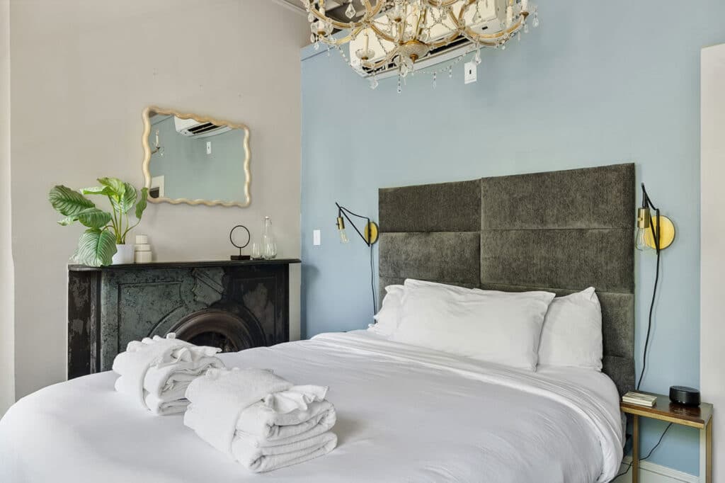 Bedroom with blue accent wall, tufted gray headboard, white bedding, chandelier, and a dark fireplace mantel.