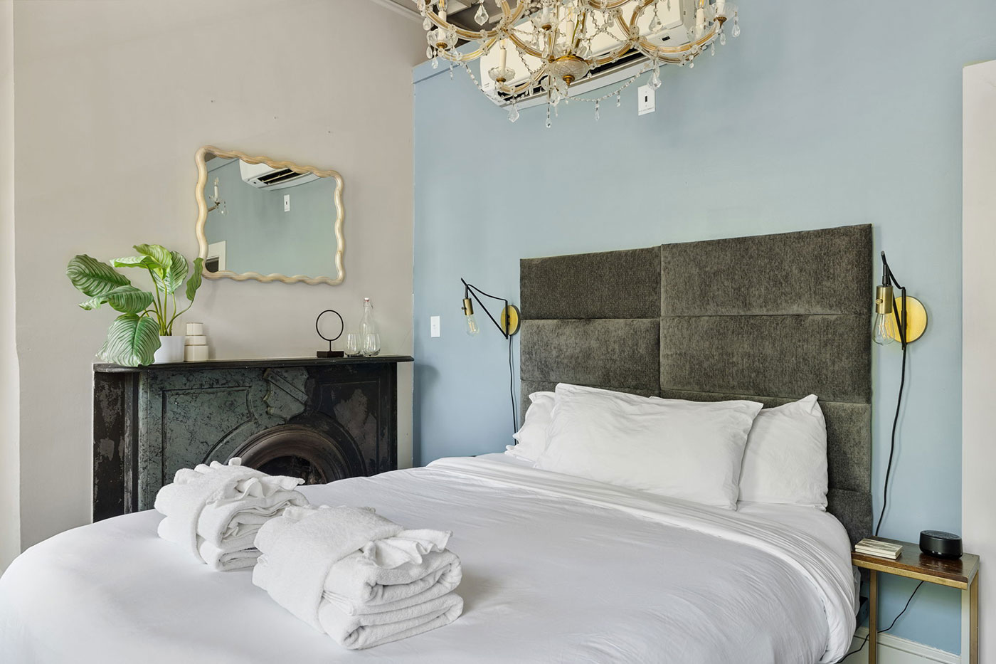 Bedroom with blue accent wall, tufted gray headboard, white bedding, chandelier, and a dark fireplace mantel.