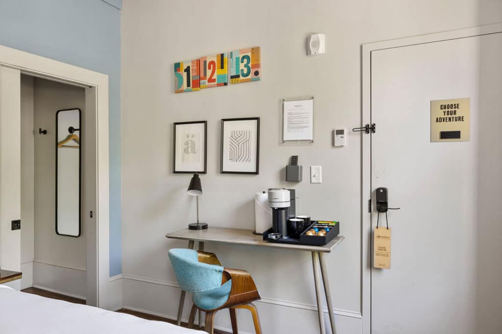 Hotel room workspace: desk with coffee station, lamp, and a teal chair against a wall with framed art.