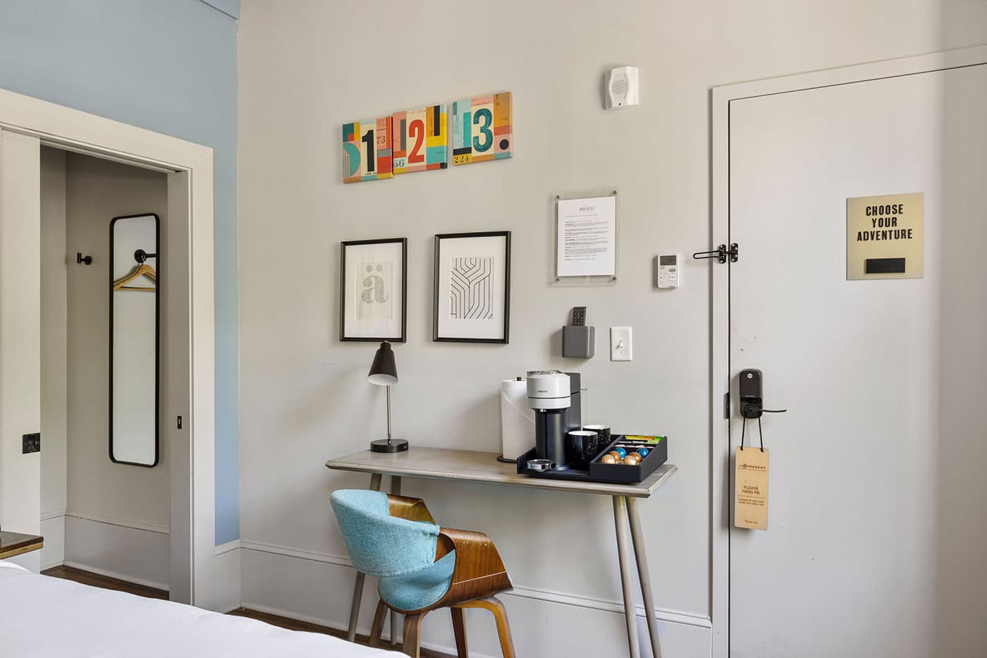 Hotel room workspace: desk with coffee station, lamp, and a teal chair against a wall with framed art.