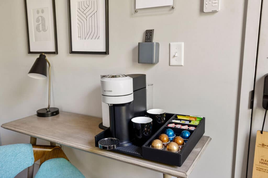 Nespresso coffee machine on a desk with a pod tray, mugs, and framed art on the wall behind it.