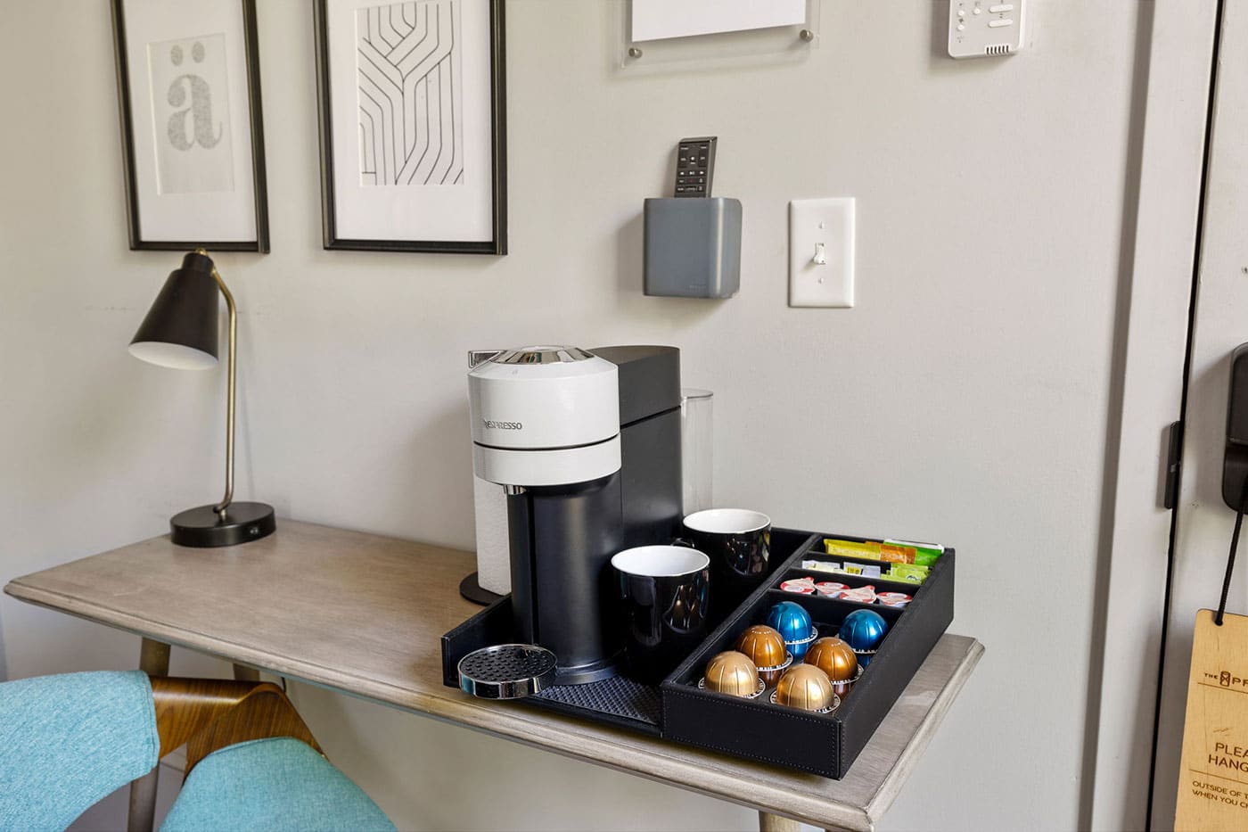Nespresso coffee machine on a desk with a pod tray, mugs, and framed art on the wall behind it.