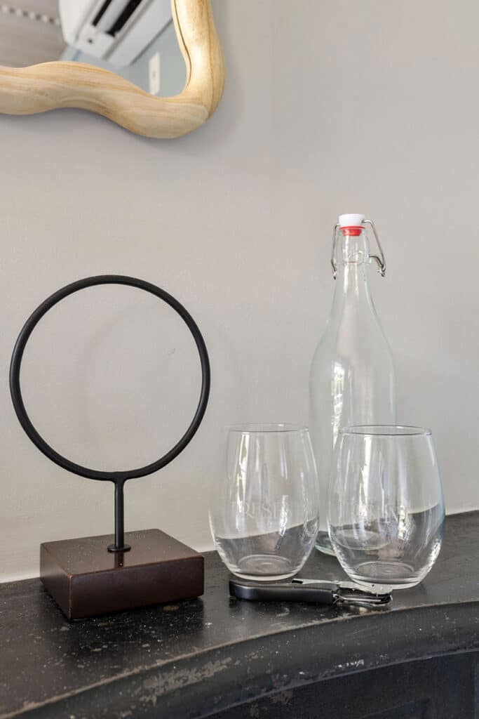 Still-life scene with a circular metal ring on a dark base, two stemless glasses, and a glass bottle with a swing-top on a black countertop.