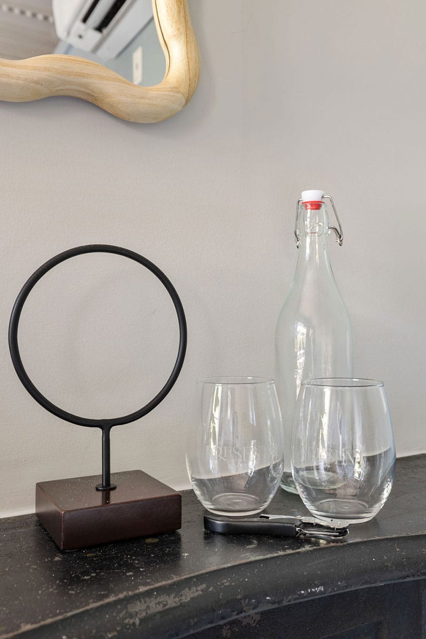 Still-life scene with a circular metal ring on a dark base, two stemless glasses, and a glass bottle with a swing-top on a black countertop.