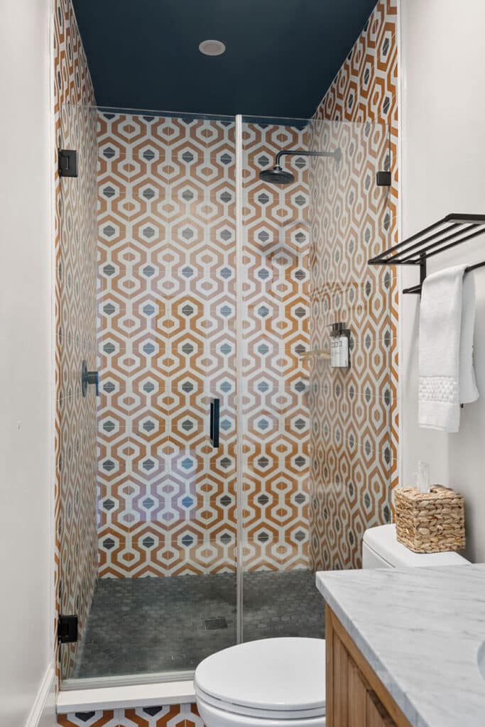Glass-walled shower with orange geometric tiles, a dark blue ceiling, and a toilet nearby beside a marble countertop.