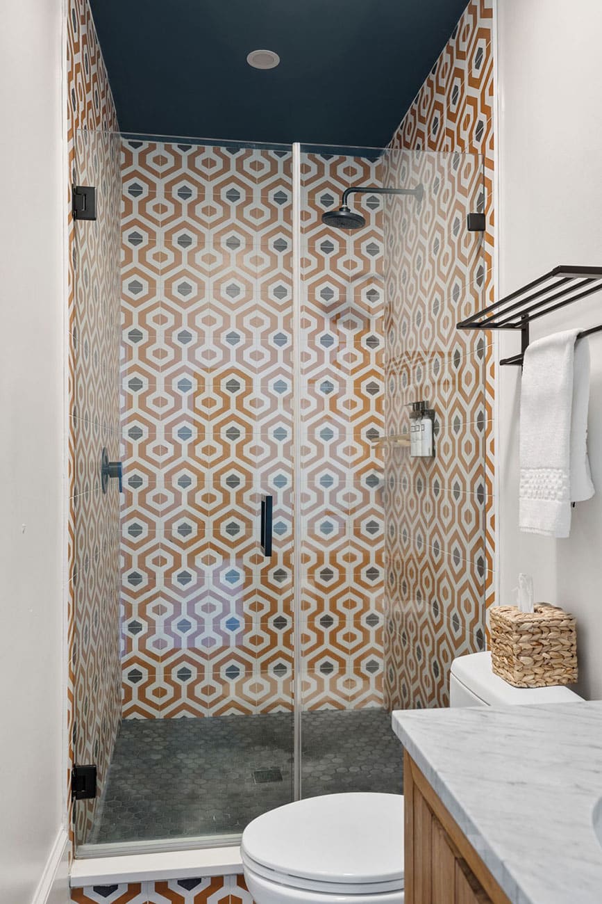 Glass-walled shower with orange geometric tiles, a dark blue ceiling, and a toilet nearby beside a marble countertop.