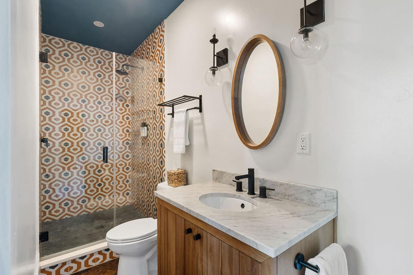 Modern bathroom with a glass shower door and bold orange geometric tile, a marble countertop vanity, and a round wooden-framed mirror.