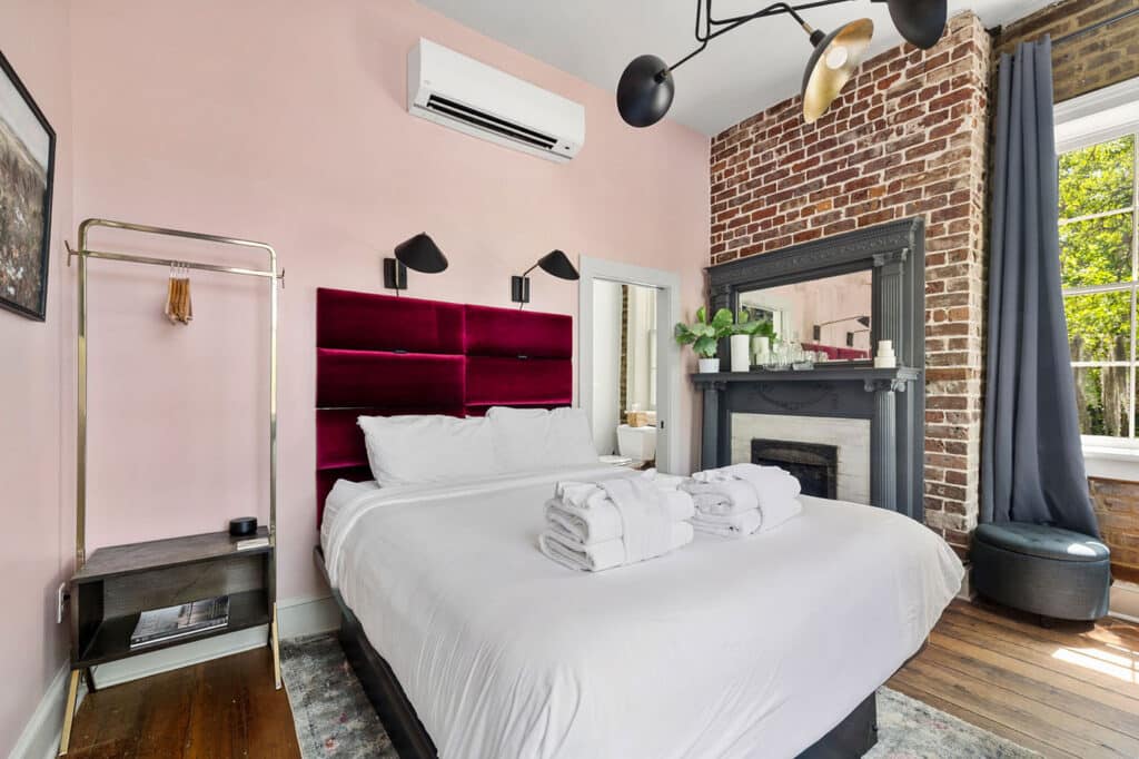 Bright hotel-style bedroom with pink walls, a burgundy tufted headboard, brick fireplace, and a large window with gray curtains.