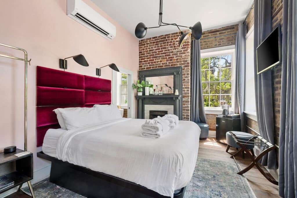 Hotel bedroom with white bedding and a bold magenta velvet headboard against a pale pink wall, brick accents, and a large window with gray curtains.