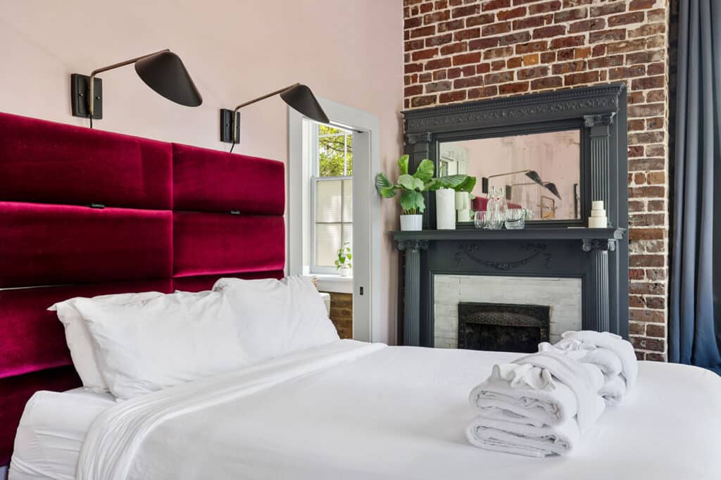 Bedroom with burgundy velvet headboard, white linens, and a brick fireplace with a dark ornate mantel and mirror on top.