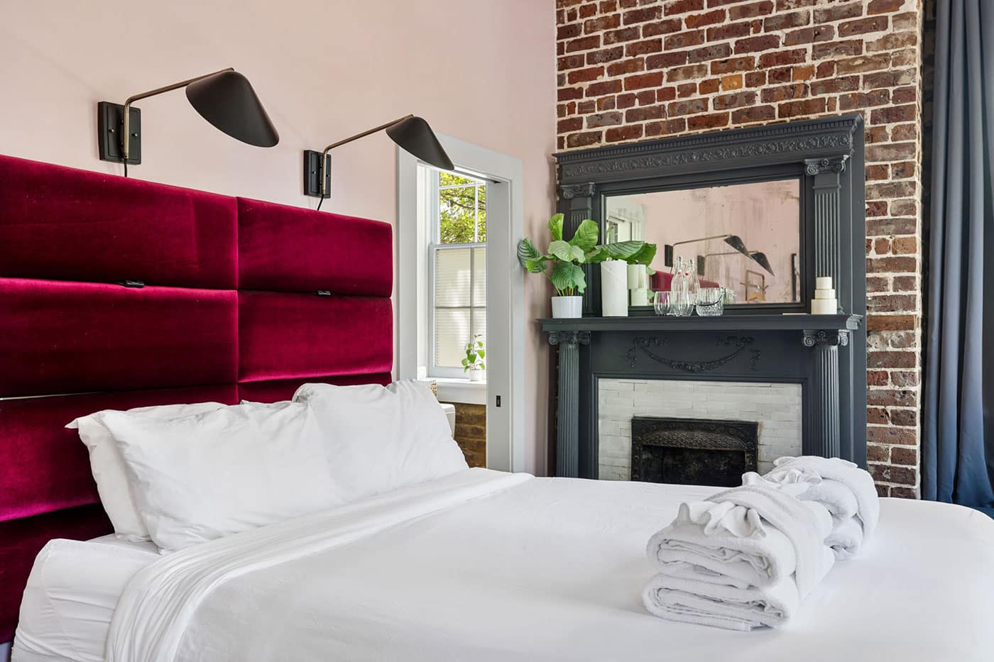 Bedroom with burgundy velvet headboard, white linens, and a brick fireplace with a dark ornate mantel and mirror on top.