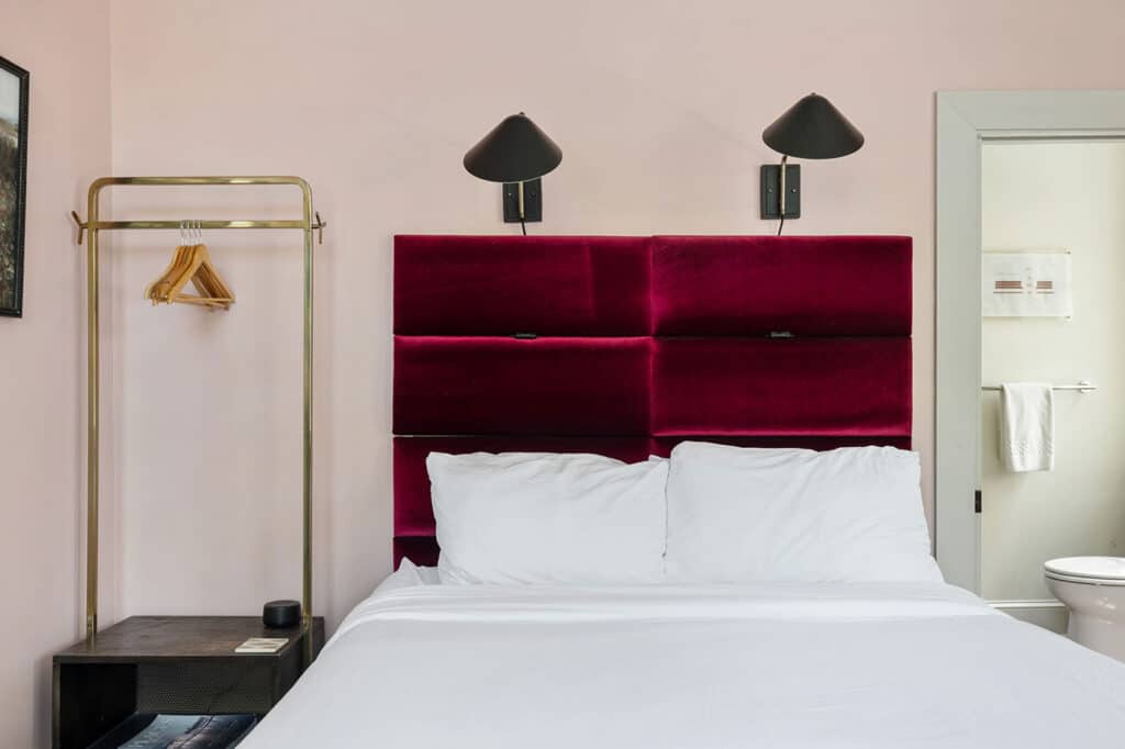 Hotel bedroom with a burgundy velvet headboard, white bedding, and black wall lamps above the bed.