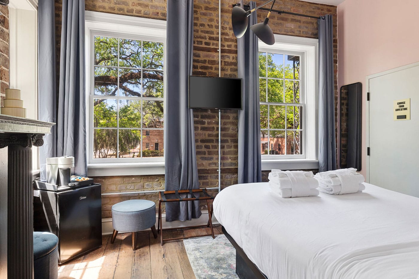 Hotel bedroom with exposed brick wall, two large windows with blue curtains, a wall-mounted TV, and a white bed with folded towels on top.