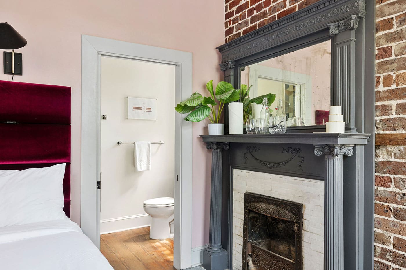 Bedroom scene with pink walls, white bed, and a burgundy headboard beside an ornate gray fireplace against a brick wall.