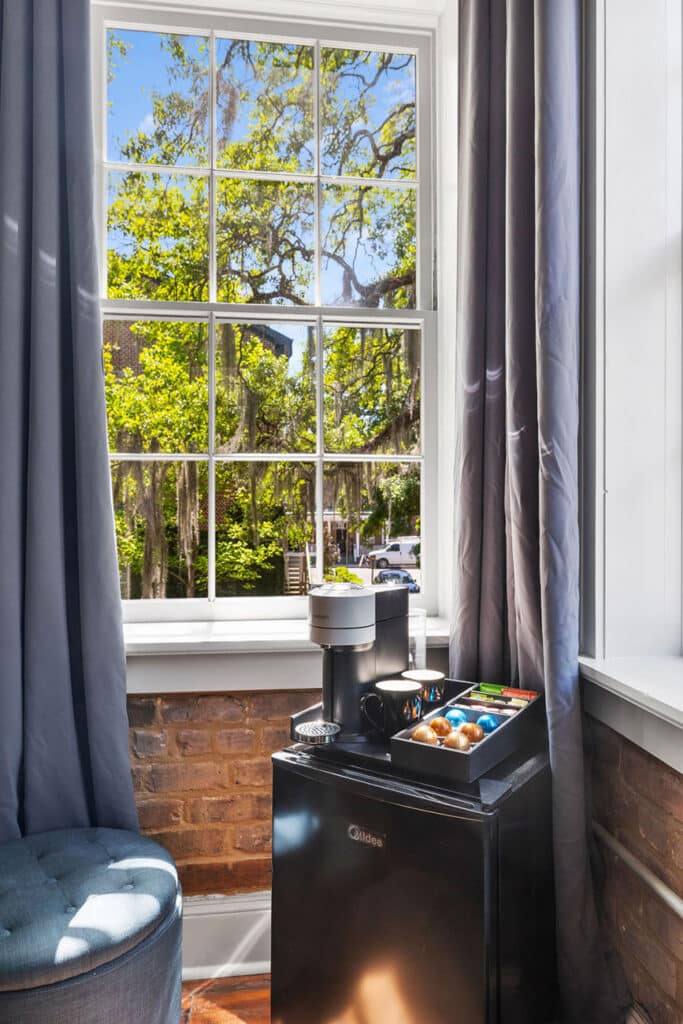Coffee station by a bright window: Nespresso machine on a small black mini fridge with mugs and a capsule tray beside gray curtains and brick walls on a sunny day.