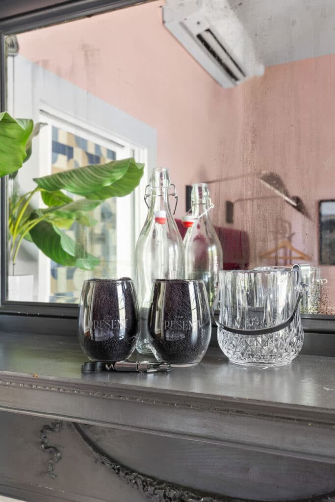 Close-up of a bar counter with two dark wine glasses, glass bottles with stoppers, and a crystal ice bucket; plant and pink wall reflected in the background