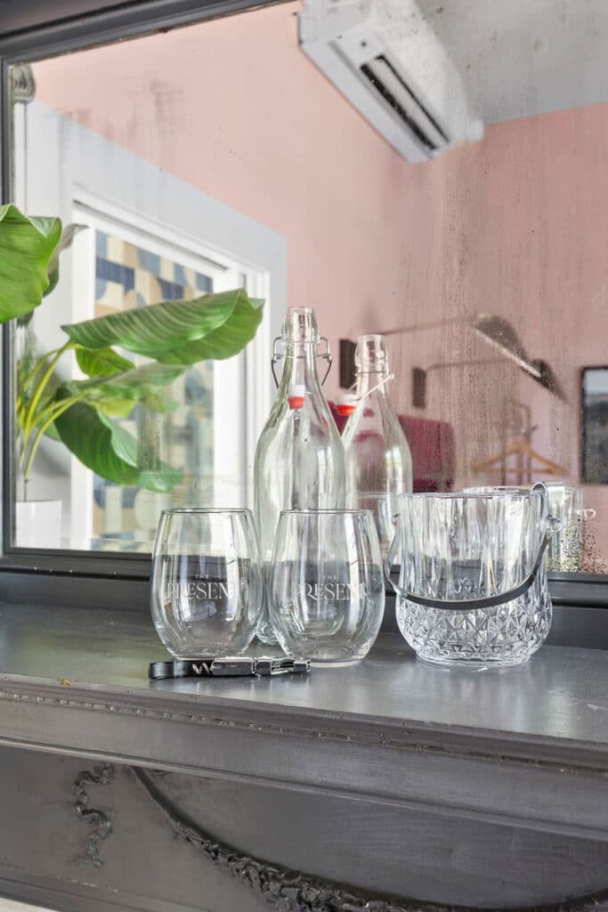 Glass bottles with swing-top caps and several clear tumblers arranged on a windowsill, with a plant in the background.