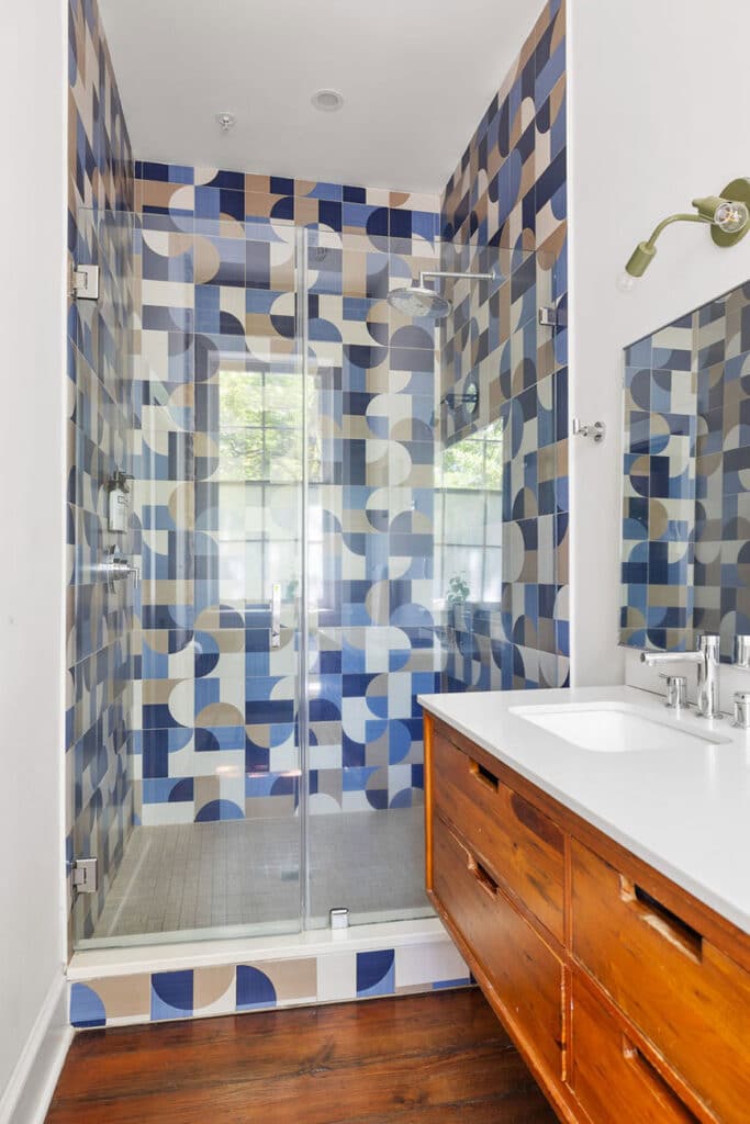 Glass-enclosed shower with blue, beige, and white patterned tile walls in a modern bathroom. Wooden vanity with white countertop and mirror to the right.