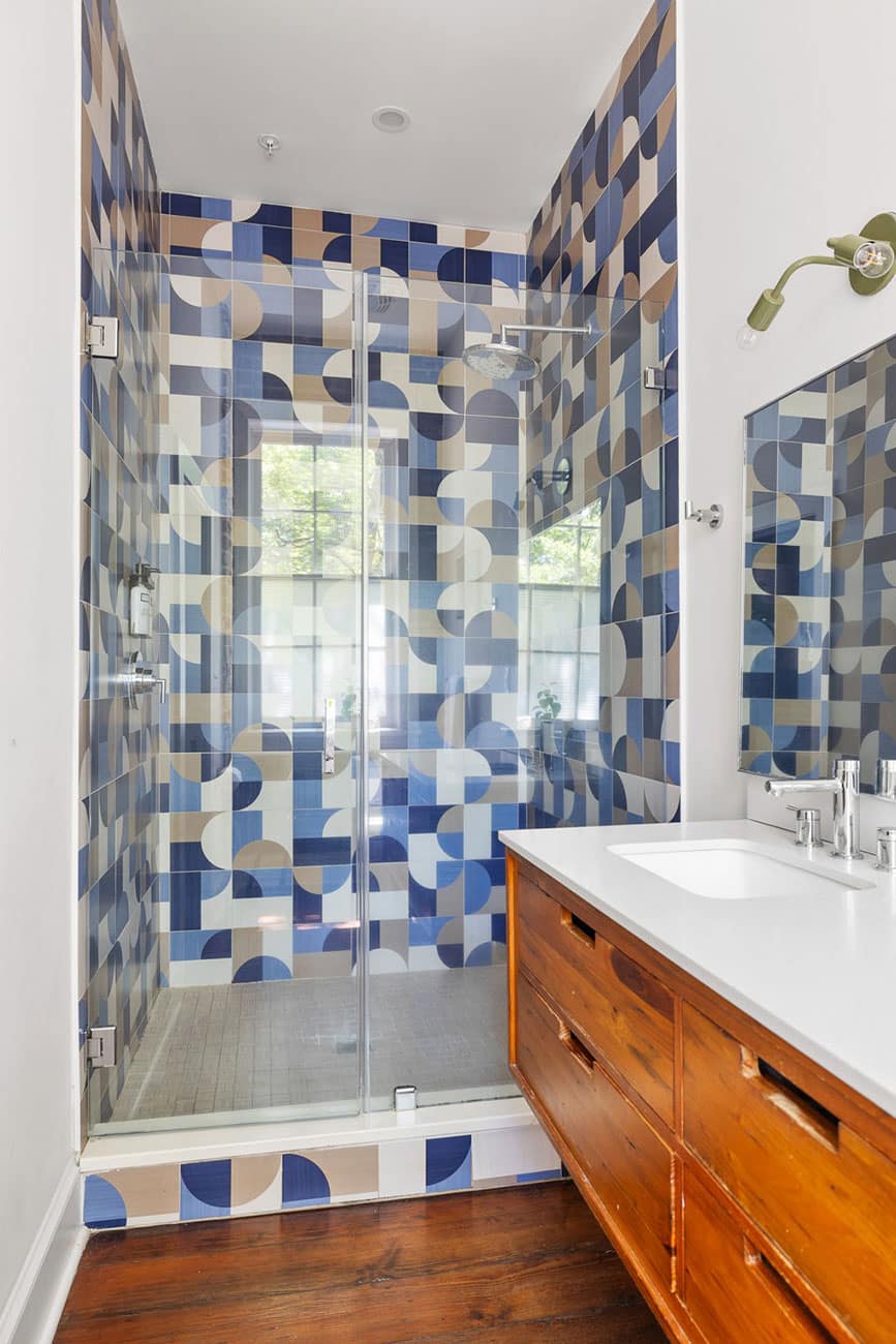 Glass-enclosed shower with blue, beige, and white patterned tile walls in a modern bathroom. Wooden vanity with white countertop and mirror to the right.