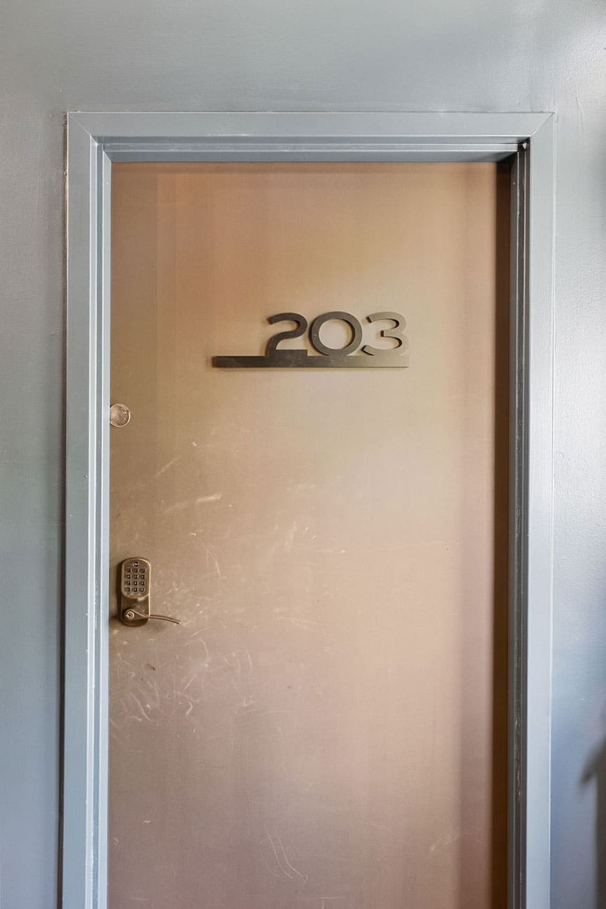Beige metal door labeled 203 with a keypad lock on a gray corridor frameLiased wall behind it