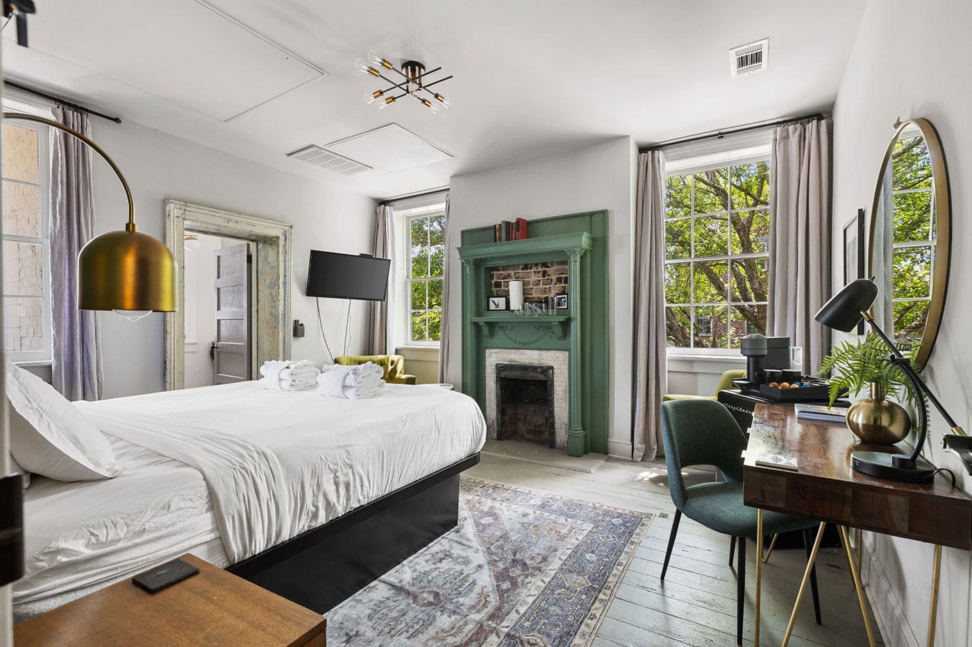Bright hotel bedroom with a white bed, gold task lamp, and a green decorative fireplace facing a rug-filled floor. Nearby desk and window seating add workspace and light.