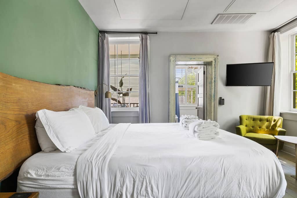 Bright hotel room with a large white bed, green accent wall, and a wooden headboard; towels on the bed and a lime green chair nearby.