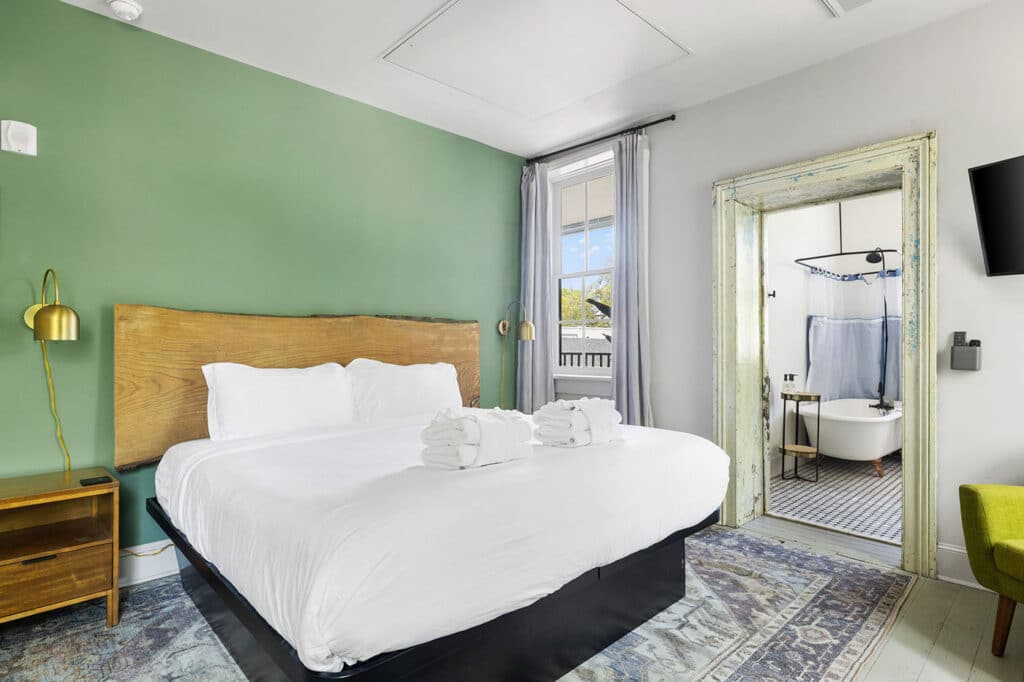 Hotel room with white bedding, green feature wall, and a doorway to a vintage-style bathroom with a freestanding tub visible through the door.