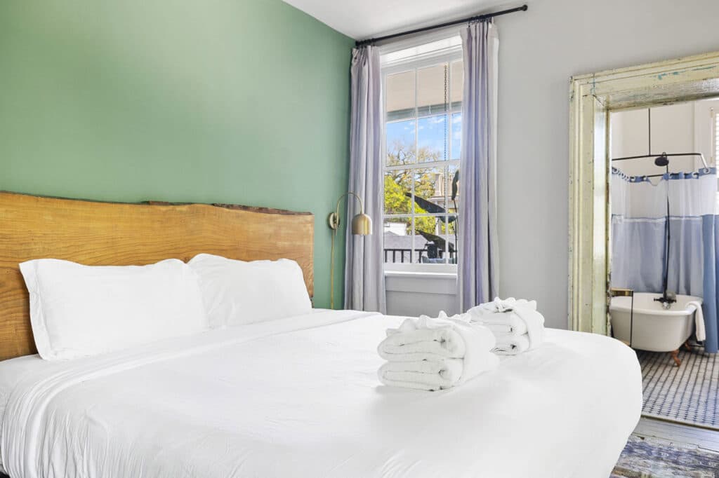 Bedroom with white bedding and a rustic wooden headboard against a green accent wall; window with lavender curtains and brass reading lamp nearby, plus a glimpse of the bathroom.