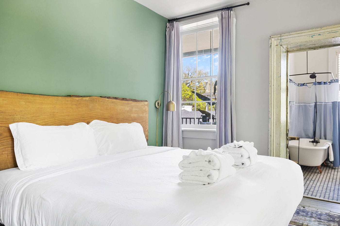 Bedroom with white bedding and a rustic wooden headboard against a green accent wall; window with lavender curtains and brass reading lamp nearby, plus a glimpse of the bathroom.
