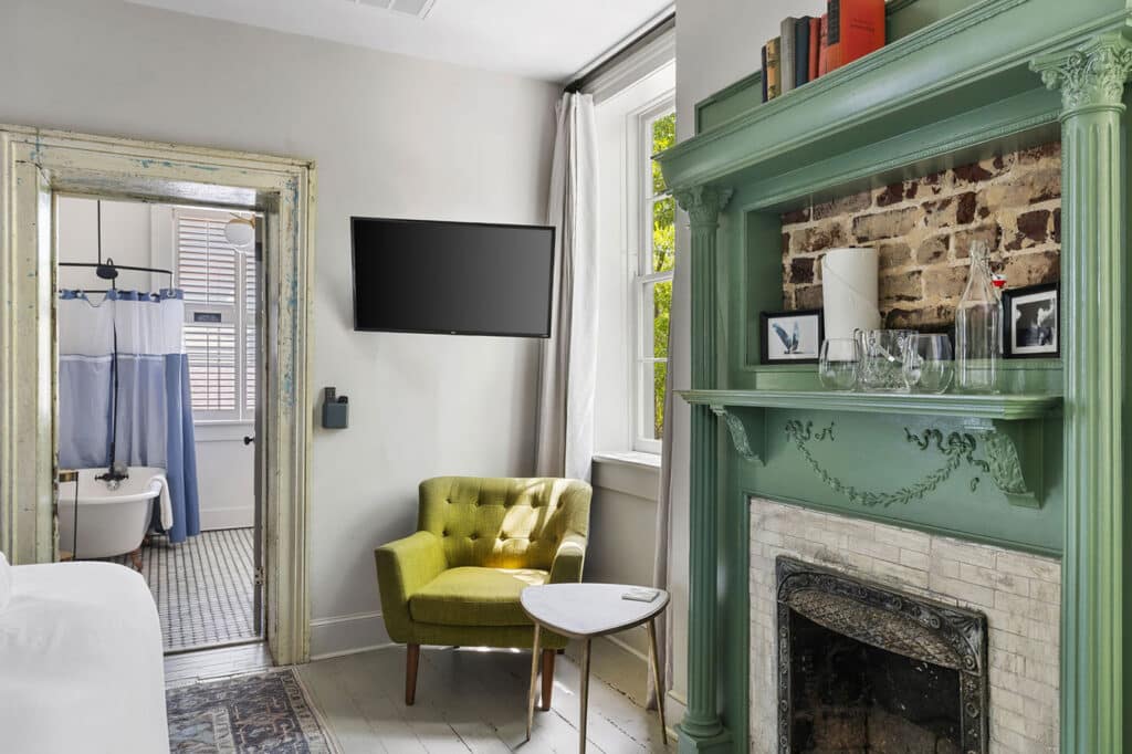 Living area of a hotel room with a green ornate fireplace, lime chair, small table, window with curtains, and a wall-mounted TV.