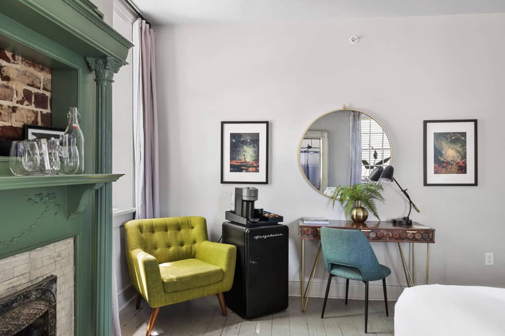Corner living space with a green fireplace mantel, mustard-yellow armchair, and a small fridge with a coffee station nearby.