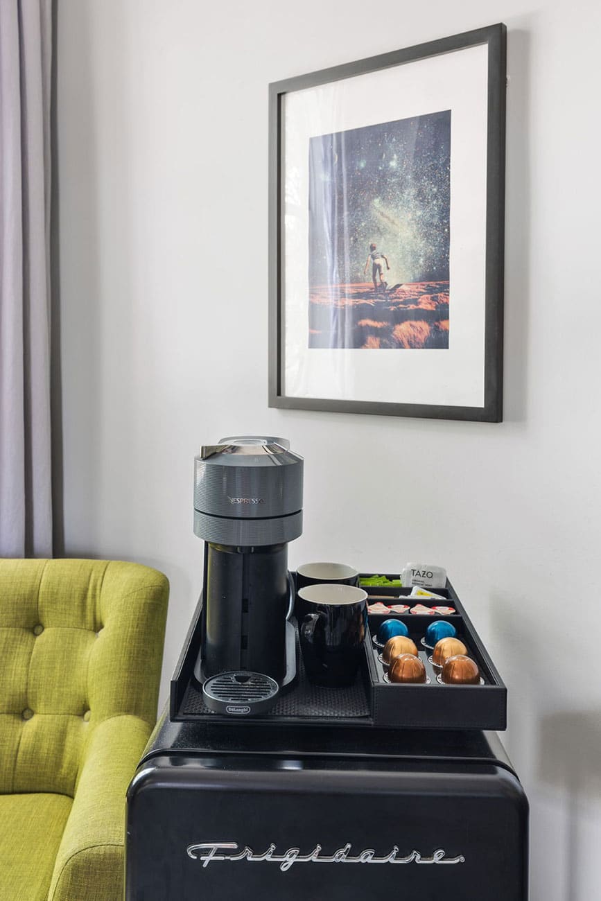 Nespresso machine on a tray with cups and coffee capsules on a small fridge, beside a lime-green chair and space-themed wall art.