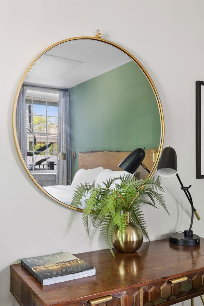Round gold-framed mirror above a wooden console table with a brass fern vase and a black desk lamp; a magazine rests on the table.